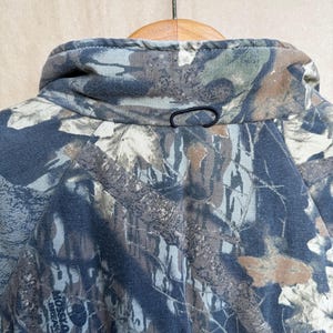 May include: Close-up of a camouflage jacket collar with a black loop closure. The jacket features a pattern of brown, green, and gray tones, typical of outdoor apparel. The collar is folded over a wooden hanger.