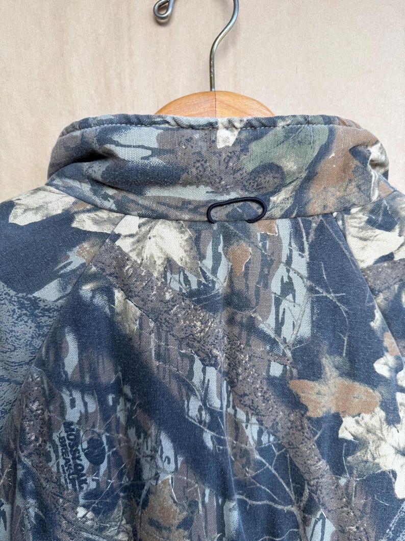May include: Close-up of a camouflage jacket collar with a black loop closure. The jacket features a pattern of brown, green, and gray tones, typical of outdoor apparel. The collar is folded over a wooden hanger.