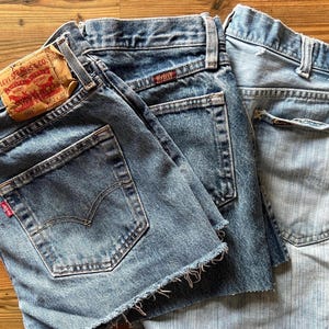 May include: Three pairs of blue denim shorts and jeans are displayed on a wooden surface. One pair of shorts has a frayed hem and a Levi Strauss & Co. label. The jeans have classic pocket designs.