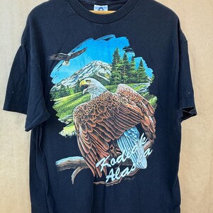 May include: Black t-shirt featuring a detailed graphic of a bald eagle perched on a branch, with a mountain and forest backdrop. The text "Kodiak Alaska" is displayed below the eagle. A second eagle is flying in the sky.