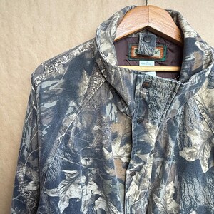 K&ouml;nnte beinhalten: Tarnjacke in Braun-, Gr&uuml;n- und Graut&ouml;nen mit Knopfleiste und hohem Kragen. Die Jacke h&auml;ngt an einem Holzb&uuml;gel. Der Stoff hat ein Bl&auml;ttermuster.