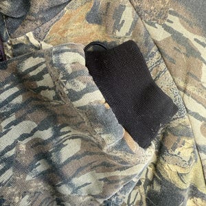 May include: Close-up of a camouflage jacket sleeve. The jacket features a pattern of brown, green, and tan hues. The sleeve has a black ribbed cuff. The fabric appears to be a durable material.