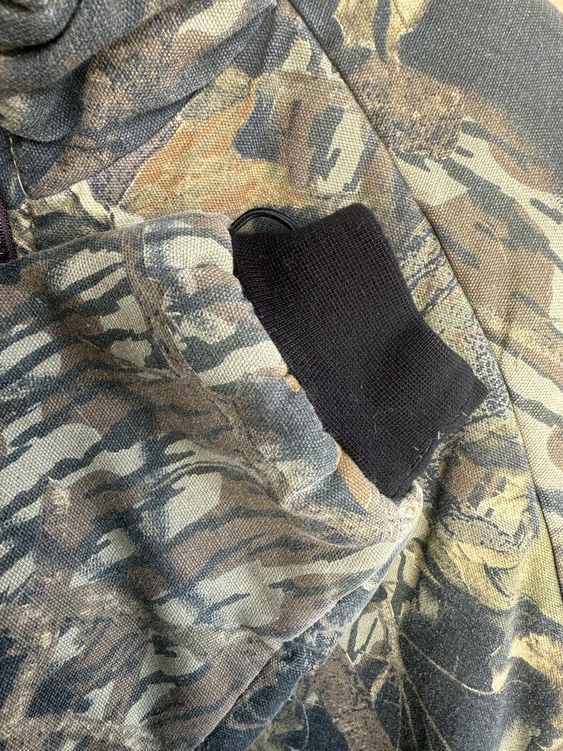 May include: Close-up of a camouflage jacket sleeve. The jacket features a pattern of brown, green, and tan hues. The sleeve has a black ribbed cuff. The fabric appears to be a durable material.