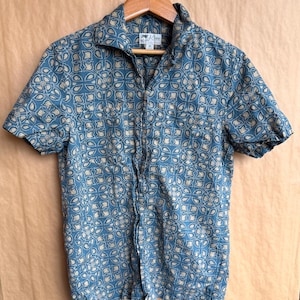 Puede incluir: Camisa azul de manga corta con un estampado floral beige repetido. La camisa tiene cuello de campamento y un pequeño bolsillo. La etiqueta de la marca "J.Crew" es visible en el cuello.