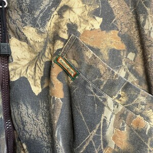 May include: Close-up of a camouflage jacket with a brown zipper and a pocket. The jacket features a pattern of brown, tan, and gray leaves. The brand name "Remington" is visible on a small tag.