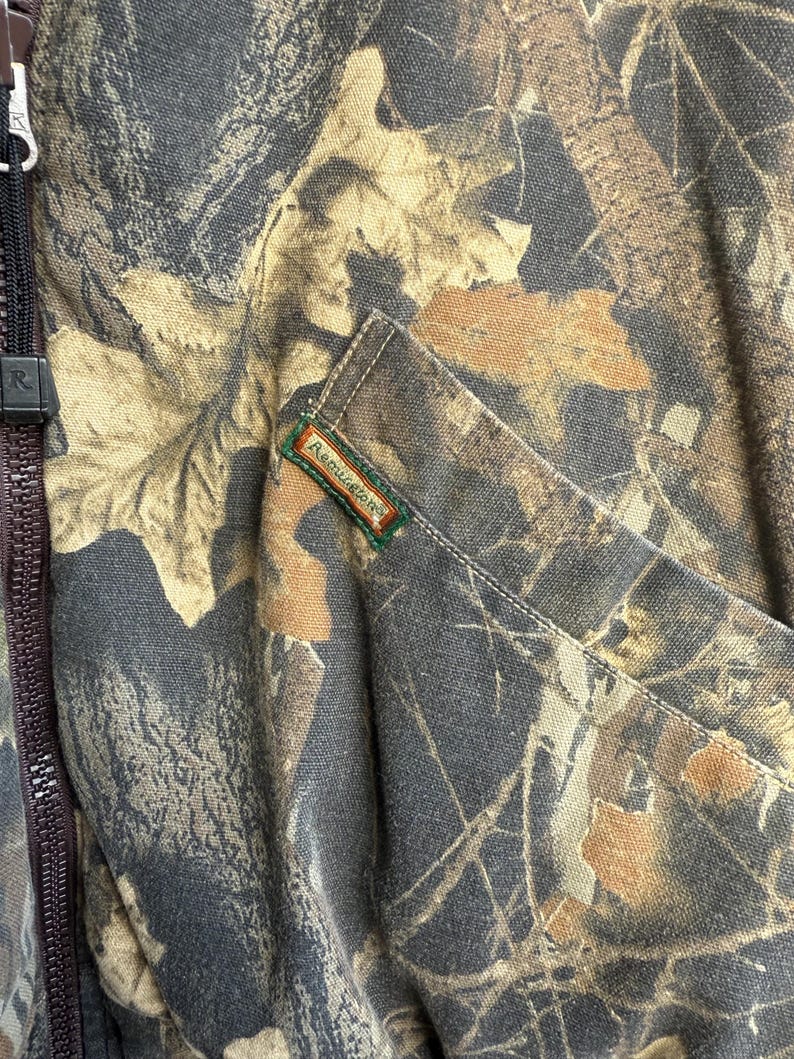 May include: Close-up of a camouflage jacket with a brown zipper and a pocket. The jacket features a pattern of brown, tan, and gray leaves. The brand name "Remington" is visible on a small tag.