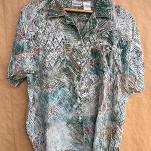 May include: A short-sleeved button-down shirt with a green, white, and coral abstract pattern. The shirt is hanging on a wooden hanger. The shirt has a collared neckline and a single chest pocket.