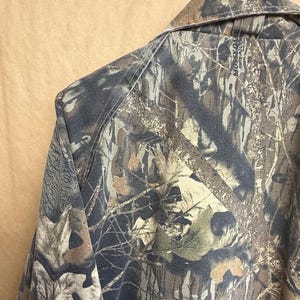 K&ouml;nnte beinhalten: Nahaufnahme eines Tarnhemdes mit einem Button-Down-Kragen. Das Hemd weist ein Muster aus braunen, gr&uuml;nen und beigen Farbt&ouml;nen auf, wobei die Worte "Mossy Oak Break-Up" auf dem Kragen zu sehen sind. Der Stoff scheint ein strapazierf&auml;higes Material zu sein.