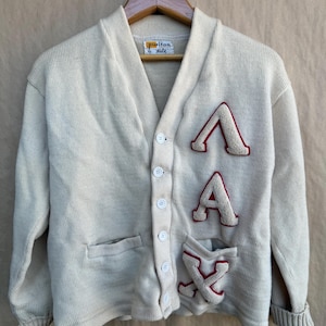 May include: Cream-colored vintage cardigan sweater with a V-neck and button closure. Features three felt letter patches in red and cream. The label reads "Puritan". The sweater has a pocket on the left side.