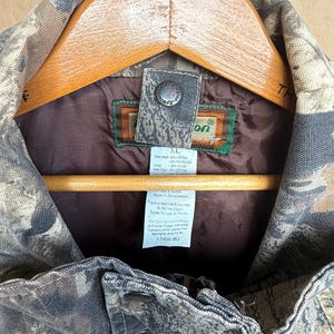 K&ouml;nnte beinhalten: Nahaufnahme einer Tarn-Jagdjacke, Gr&ouml;&szlig;e XL, an einem Holzb&uuml;gel. Die Jacke hat ein braunes Futter und einen Knopfverschluss. Das Etikett gibt an, dass die Au&szlig;enschale aus 60 % Baumwolle und 40 % Polyester besteht.