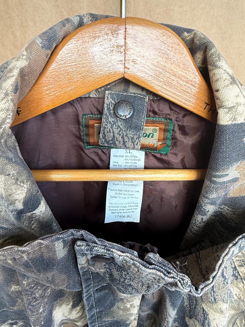 K&ouml;nnte beinhalten: Nahaufnahme einer Tarn-Jagdjacke, Gr&ouml;&szlig;e XL, an einem Holzb&uuml;gel. Die Jacke hat ein braunes Futter und einen Knopfverschluss. Das Etikett gibt an, dass die Au&szlig;enschale aus 60 % Baumwolle und 40 % Polyester besteht.