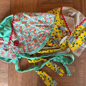 May include: Vintage baby bonnet in mint green, floral, and yellow patterns. The bonnet features red trim, a red button, and ties. The floral pattern includes red flowers on a light blue background. The yellow pattern has blue birds.