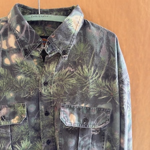 May include: A button-down shirt with a camouflage pattern featuring green and gray pine needles. The shirt has two chest pockets and black buttons. The collar has the text "Ernie D. Lewis".