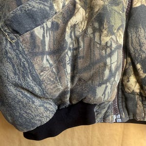 May include: Close-up of a camouflage jacket with a black ribbed cuff. The jacket features a zipper closure and a pattern of brown, green, and gray tones, typical of outdoor wear.