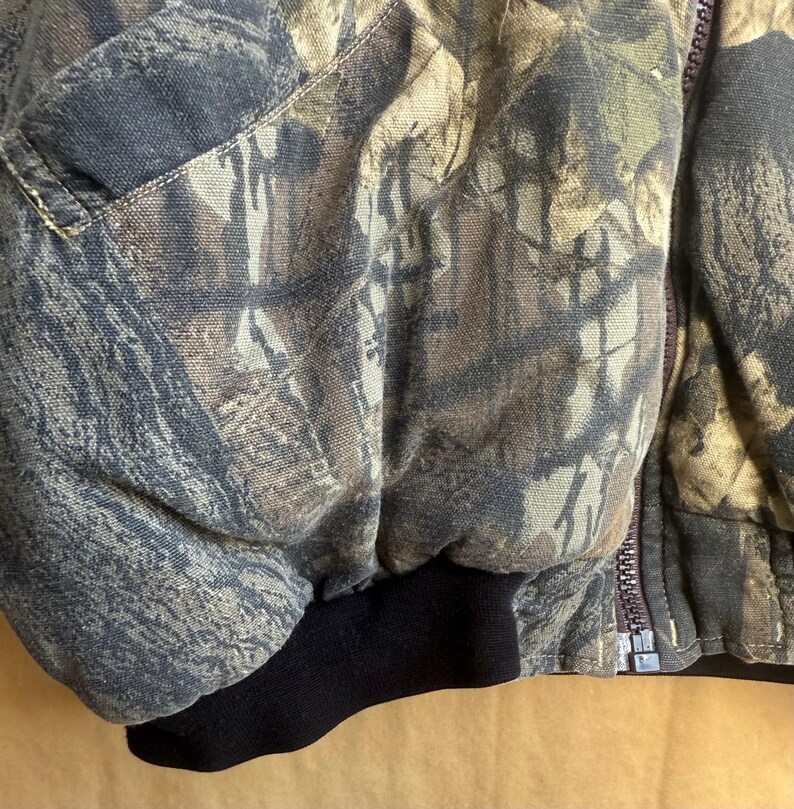 May include: Close-up of a camouflage jacket with a black ribbed cuff. The jacket features a zipper closure and a pattern of brown, green, and gray tones, typical of outdoor wear.