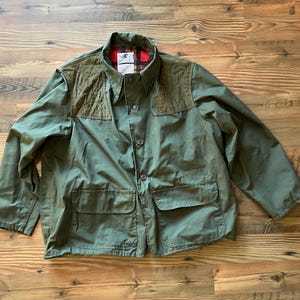 May include: Olive green jacket with a button-up front, two front pockets, and brown shoulder patches. The collar and inner lining feature a red and black plaid pattern. A classic, durable outerwear piece.