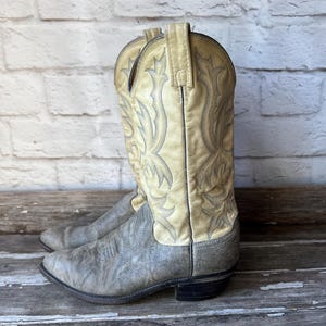 May include: A pair of worn cowboy boots. The boots are two-toned, with a light tan upper and a gray lower section. Intricate stitching in a darker color adorns the upper. The boots have a classic Western design.