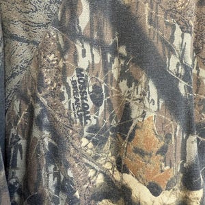 May include: Close-up of a camouflage fabric with a blend of brown, green, and tan hues. The fabric features a detailed pattern of leaves and branches, with the text "MOSSY OAK BREAK-UP" visible.