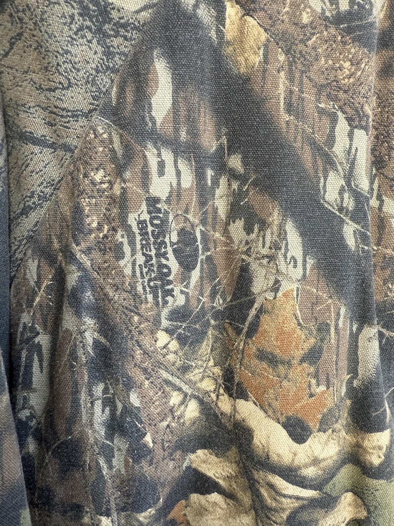 May include: Close-up of a camouflage fabric with a blend of brown, green, and tan hues. The fabric features a detailed pattern of leaves and branches, with the text "MOSSY OAK BREAK-UP" visible.