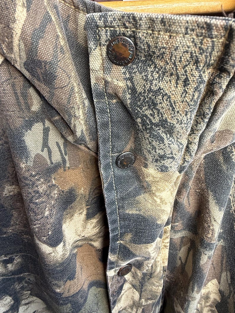 K&ouml;nnte beinhalten: Nahaufnahme einer Tarnjacke mit drei sichtbaren Metallkn&ouml;pfen. Die Jacke weist ein Muster aus Braun, Beige und Schwarz auf, mit einem Knopfverschluss. Der Stoff scheint ein strapazierf&auml;higes, m&ouml;glicherweise Canvas-Material zu sein.