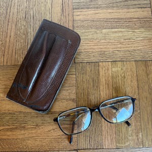 May include: Eyeglasses with a brown tortoiseshell frame and clear lenses, next to a brown leather glasses case. The glasses have a rectangular shape and thin metal accents. The case is rectangular with a textured surface.