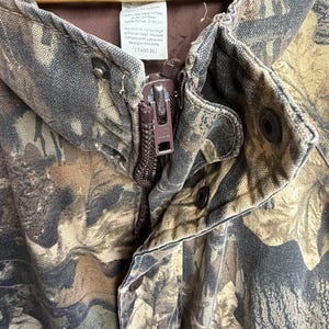 K&ouml;nnte beinhalten: Nahaufnahme einer Tarnjacke mit braunem Rei&szlig;verschluss und Druckkn&ouml;pfen. Die Jacke hat ein braunes Innenfutter und ein Etikett mit der Gr&ouml;&szlig;e XL. Die Jacke ist aus strapazierf&auml;higem Stoff im Outdoor-Stil gefertigt.