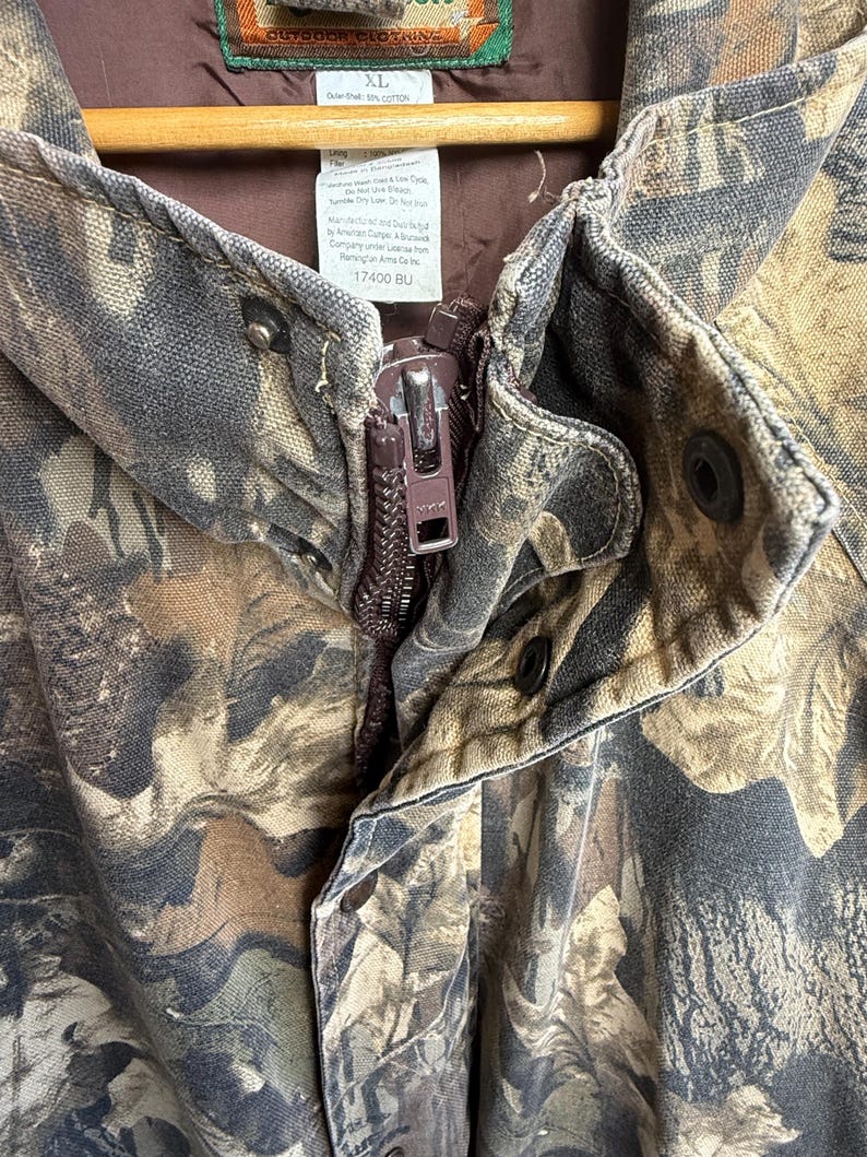 K&ouml;nnte beinhalten: Nahaufnahme einer Tarnjacke mit braunem Rei&szlig;verschluss und Druckkn&ouml;pfen. Die Jacke hat ein braunes Innenfutter und ein Etikett mit der Gr&ouml;&szlig;e XL. Die Jacke ist aus strapazierf&auml;higem Stoff im Outdoor-Stil gefertigt.