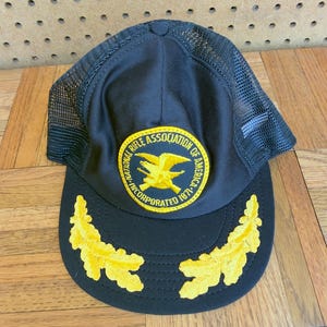 May include: A black trucker hat featuring a circular patch with a yellow eagle emblem and the text National Rifle Association of America Incorporated 1871. The brim is decorated with two yellow embroidered oak leaf clusters on each side.