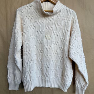 May include: Cream-colored knit sweater with a mock turtleneck and textured, raised floral pattern. The long-sleeved sweater is hanging on a wooden hanger against a neutral background. Ribbed cuffs and hem.