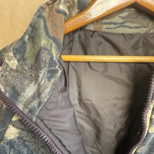 May include: Close-up of a camouflage jacket with a full zipper. The jacket features a woodland camouflage pattern in shades of green, brown, and black. The interior lining is a solid dark brown color. The jacket is hanging on a wooden hanger.