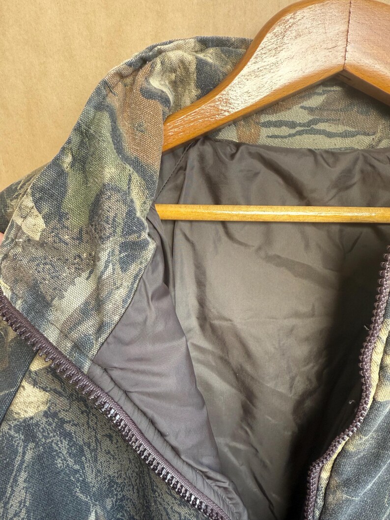 May include: Close-up of a camouflage jacket with a full zipper. The jacket features a woodland camouflage pattern in shades of green, brown, and black. The interior lining is a solid dark brown color. The jacket is hanging on a wooden hanger.