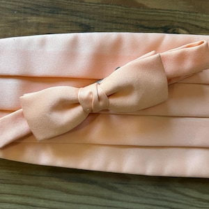 May include: A peach-colored cummerbund and bow tie set. The cummerbund is pleated and the bow tie is pre-tied. The set is made of a smooth, satin-like material. The bow tie is attached to the cummerbund with a small, black clasp.