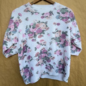 May include: A white short-sleeved sweater featuring a textured knit pattern and a floral print in shades of pink, purple, and muted green. The garment has a ribbed crew neckline and ribbed cuffs on the sleeves and hem, providing a structured fit.