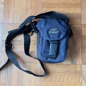 May include: A navy blue Tamrac Digital Series camera bag with a black adjustable shoulder strap. The bag features a front flap secured by a black plastic buckle and a small oval patch with the text Tamrac Digital Series on the front.