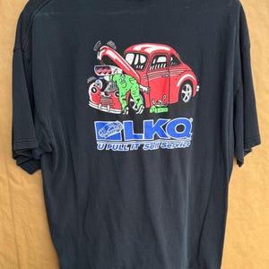 Puede incluir: Camiseta negra con un gráfico de un coche rojo siendo reparado por una figura verde con aspecto de alienígena. El texto "LKQ U Pull It Self Service" está impreso debajo en azul y blanco.