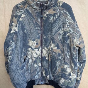 May include: A full-zip camouflage jacket with a high collar and two front pockets. The jacket features a brown zipper and a black ribbed hem and cuffs. The camouflage pattern includes shades of green, brown, and gray.