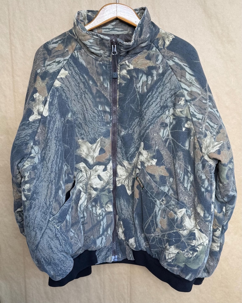May include: A full-zip camouflage jacket with a high collar and two front pockets. The jacket features a brown zipper and a black ribbed hem and cuffs. The camouflage pattern includes shades of green, brown, and gray.