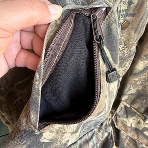 K&ouml;nnte beinhalten: Nahaufnahme einer Tarnjacke mit Rei&szlig;verschlusstasche. Die Tasche ist ge&ouml;ffnet und zeigt ein schwarzes Innenfutter. Die Jacke hat einen braunen Rei&szlig;verschluss und eine schwarze Kordel. Der Stoff hat ein braunes, gr&uuml;nes und beiges Muster.