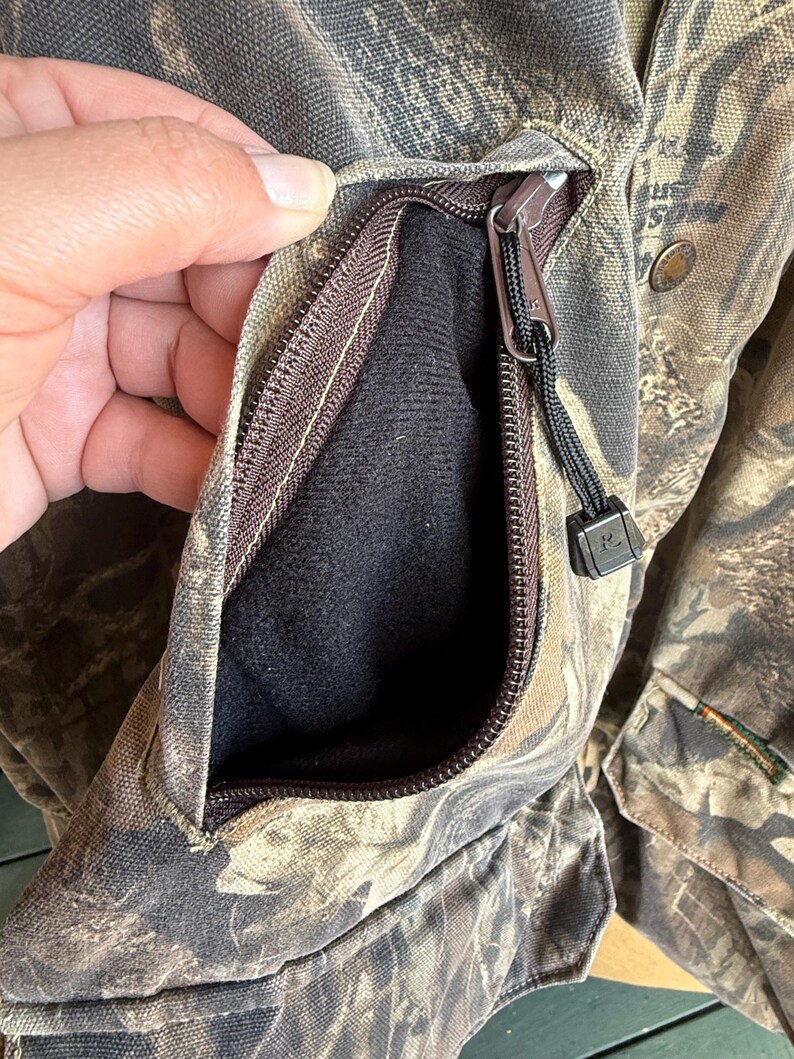 K&ouml;nnte beinhalten: Nahaufnahme einer Tarnjacke mit Rei&szlig;verschlusstasche. Die Tasche ist ge&ouml;ffnet und zeigt ein schwarzes Innenfutter. Die Jacke hat einen braunen Rei&szlig;verschluss und eine schwarze Kordel. Der Stoff hat ein braunes, gr&uuml;nes und beiges Muster.