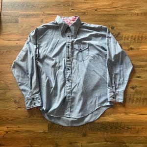 May include: A long-sleeve button-down shirt in light blue with a pointed collar. The shirt features a single chest pocket, dark buttons, and a red and white gingham check pattern visible on the inner collar and the underside of the cuffs.