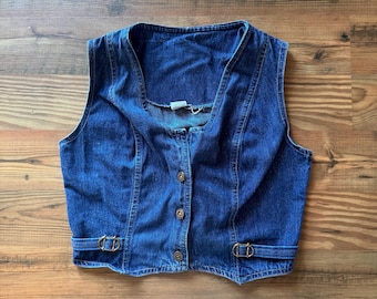Op de afbeelding: Een donkerblauw spijkergilet met een diepe V-hals, een knoopsluiting aan de voorzijde met drie metalen knopen en twee verstelbare zijbanden met goudkleurige metalen gespen in de taille. Het kledingstuk is voorzien van zichtbare naden.