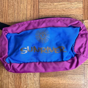 May include: A rectangular fanny pack featuring a vibrant blue center panel and purple borders. The blue section displays a gold sunburst icon above the text SUNRIVER in gold lettering. A black strap is attached to the side of the zippered bag.