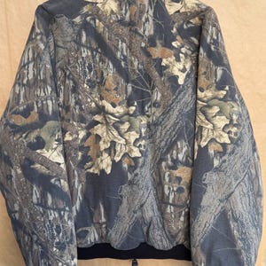 May include: A camouflage jacket with a full zip closure and a black ribbed hem and cuffs. The jacket features a pattern of brown, tan, and green leaves and branches on a dark gray background. The jacket has a high collar.