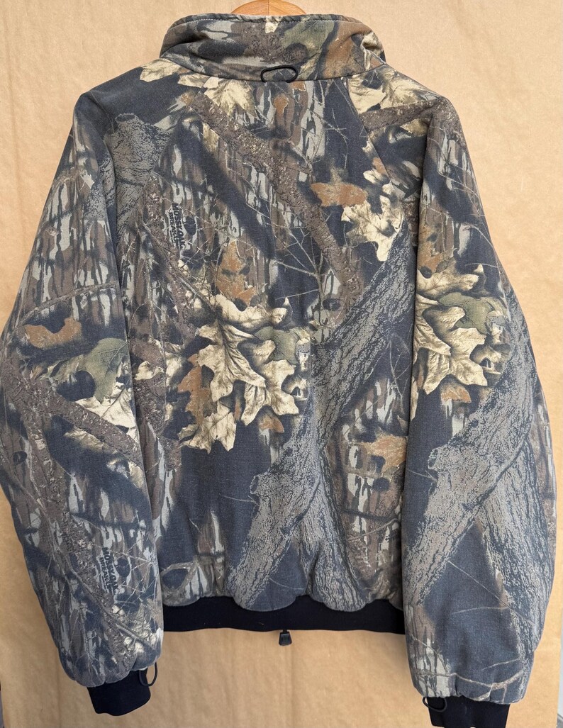 May include: A camouflage jacket with a full zip closure and a black ribbed hem and cuffs. The jacket features a pattern of brown, tan, and green leaves and branches on a dark gray background. The jacket has a high collar.