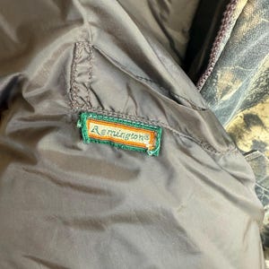 May include: Close-up of a brown jacket interior with a green, orange, and white embroidered "Remington" patch. The jacket has a smooth, slightly shiny texture. A camouflage pattern is visible in the background.