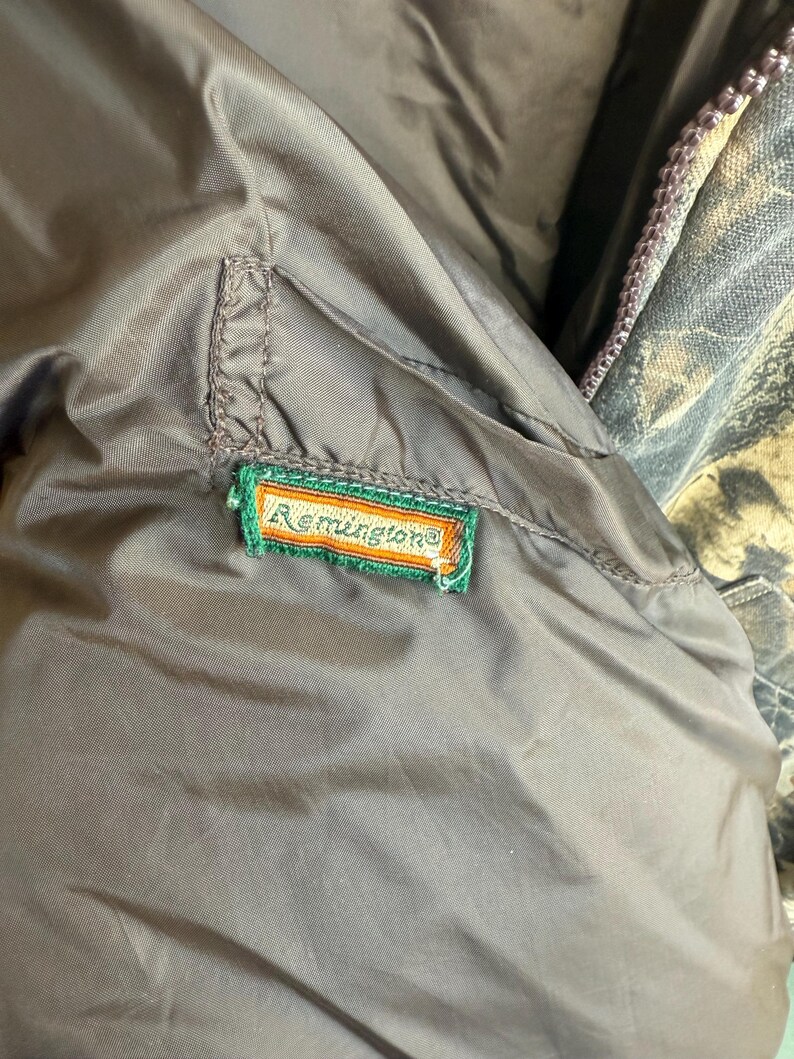 May include: Close-up of a brown jacket interior with a green, orange, and white embroidered "Remington" patch. The jacket has a smooth, slightly shiny texture. A camouflage pattern is visible in the background.