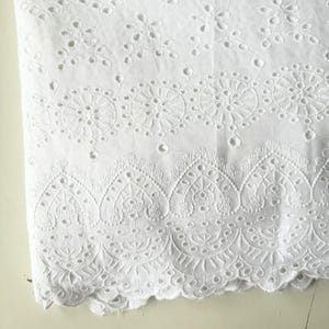 May include: Close-up of white eyelet fabric. The fabric features intricate floral and geometric cut-out patterns. The bottom edge has a scalloped design. The fabric is likely cotton or a cotton blend.