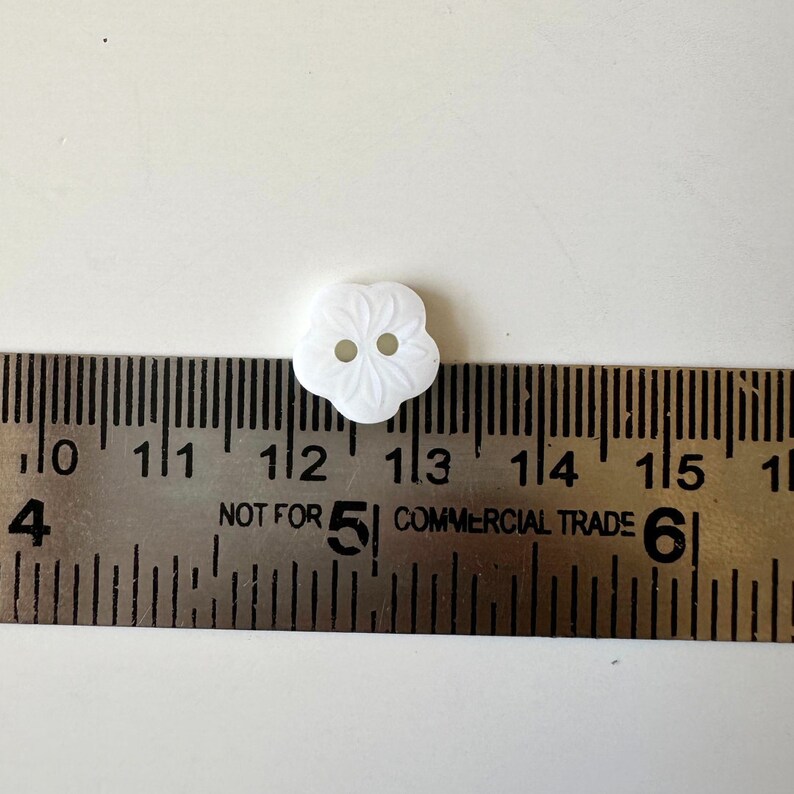 K&ouml;nnte beinhalten: Ein wei&szlig;er Plastikknopf mit Blumenmuster. Der Knopf hat einen Durchmesser von etwa 3,2 cm.