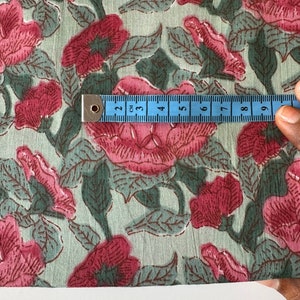 May include: Close-up of a light green fabric with a pink floral print. A blue measuring tape is placed on the fabric, showing 9 centimeters.