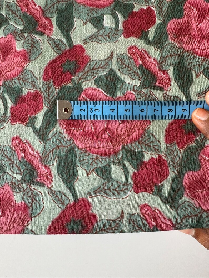 May include: Close-up of a light green fabric with a pink floral print. A blue measuring tape is placed on the fabric, showing 9 centimeters.